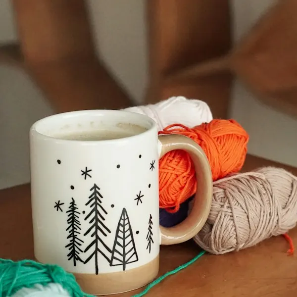 A white mug with painted fir trees beside balls of yarn