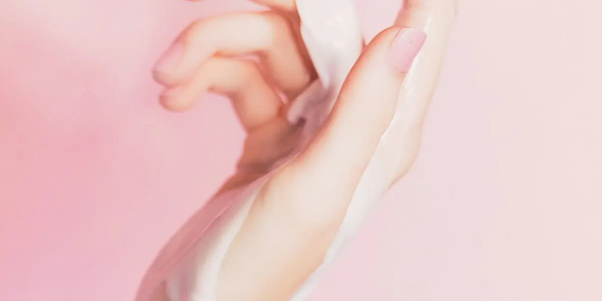 A hand with a swirl of white cream on a pink background